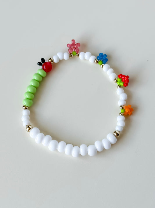 A bracelet with white beads and a caterpillar-shaped charm, featuring colorful beaded fruit.